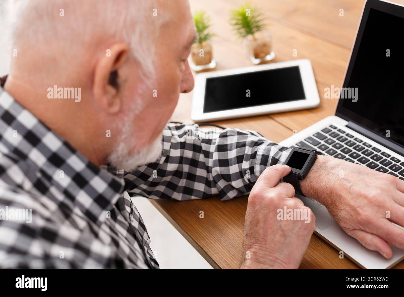 Senior Man Checking Smartwatch While Working on Laptop at Home Office Stock Photo