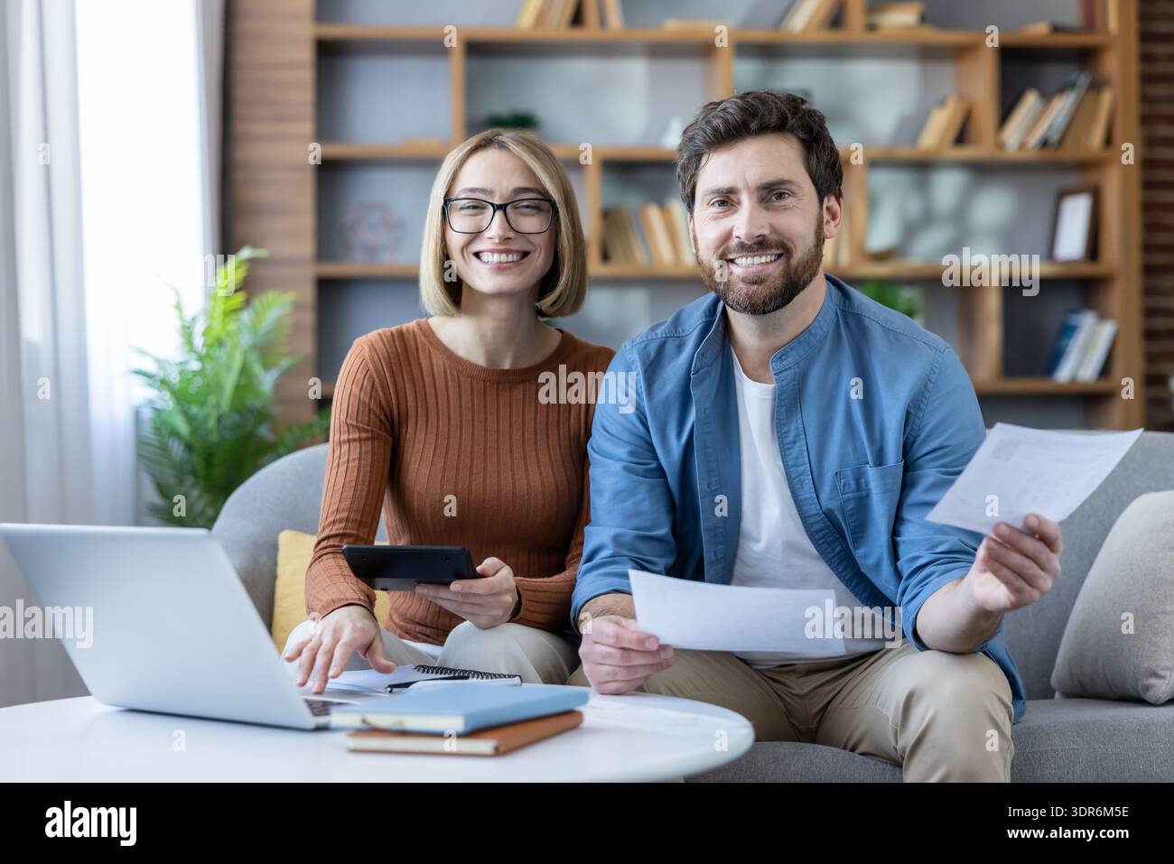 Smiling young couple sitting on a couch at home, collaborating on personal finances, calculating expenses, and filing taxes using a laptop and documen Stock Photo