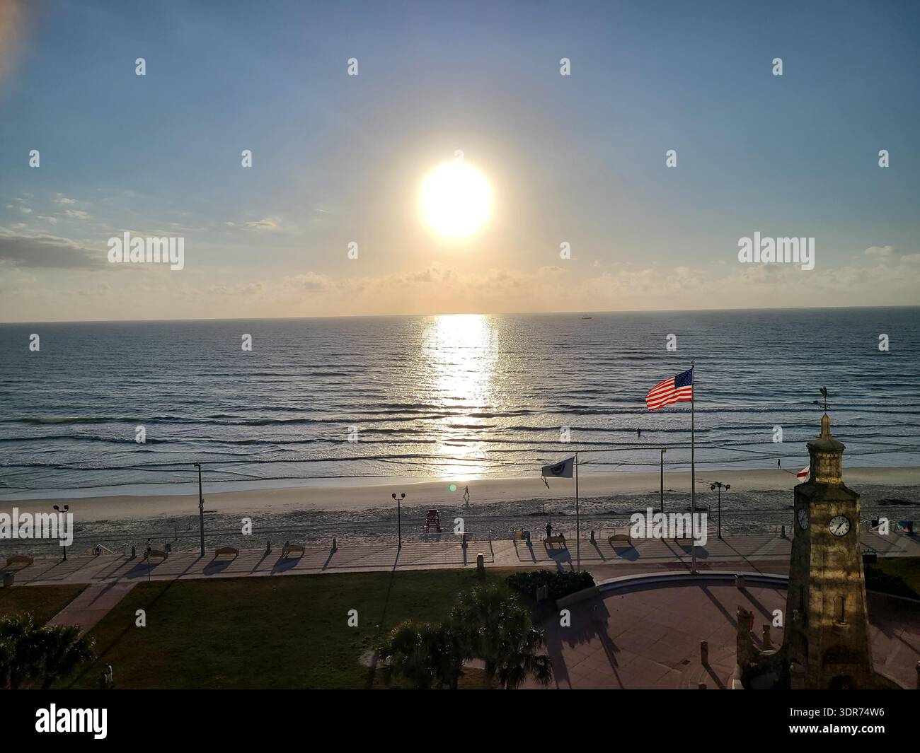 EDITORIAL USE ONLY- DAYTONA BEACH FLORIDA:  ​A stunning elevated perspective of the Atlantic coastline at Daytona Beach, Florida, featuring the iconic Coquina Clock Tower. The sun hangs low and bright in a clear blue sky, creating a brilliant golden reflection on the ocean surface. An American flag ripples in the sea breeze in the foreground, framing a peaceful scene of the beach, boardwalk, and the historic stone masonry of the clock tower during the golden hour. - Smartphone Captured Stock Image