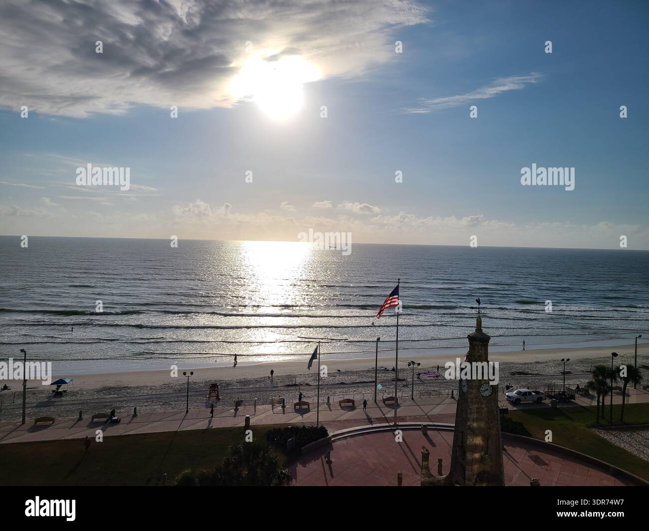 EDITORIAL USE ONLY- DAYTONA BEACH FLORIDA:  ​A high-angle, scenic view of the historic Coquina Clock Tower at the Daytona Beach Boardwalk in Florida, USA. The bright morning sun is centered in a hazy, cloud-streaked sky, casting a brilliant shimmering reflection across the gentle waves of the Atlantic Ocean. In the foreground, an American flag ripples in the coastal breeze next to the stone tower, overlooking a wide expanse of white sand beach and a quiet pedestrian promenade as the day begins. - Smartphone Captured Stock Image