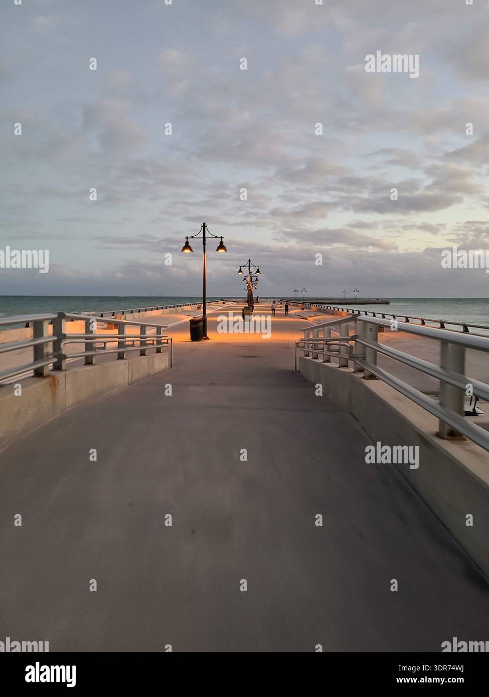 EDITORIAL USE ONLY- KEY WEST FLORIDA:  A wide-angle, symmetrical perspective of the Edward B. Knight Pier (White Street Pier) during twilight in Key West, Florida. The concrete promenade is illuminated by warm lamp posts, casting orange light on the ground against a backdrop of soft blue evening clouds and the Atlantic Ocean. Two distant figures are seen walking along the vast, peaceful structure. - Smartphone Captured Stock Image