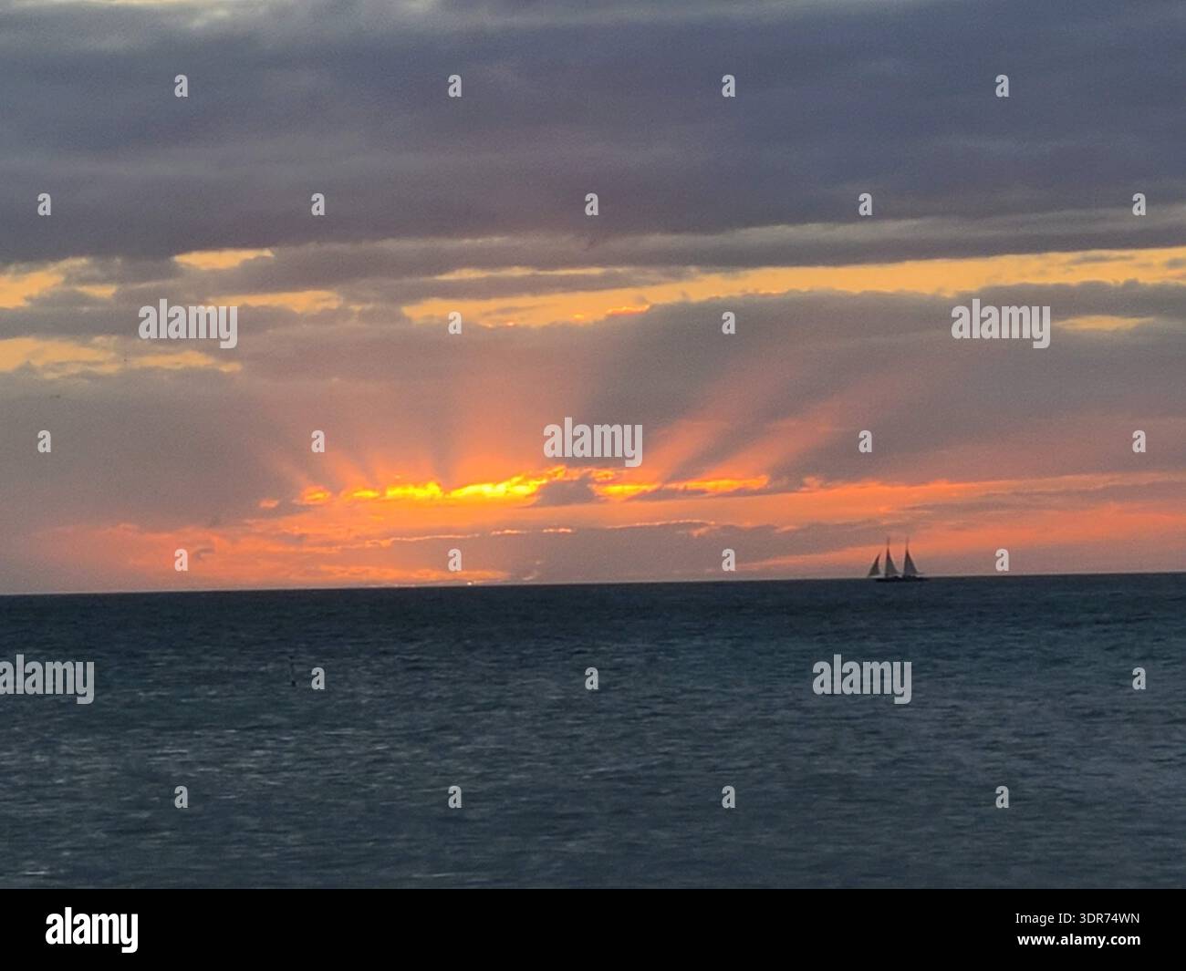 EDITORIAL USE ONLY- KEY WEST FLORIDA:  Scenic coastal landscape at dusk in Key West, Florida, featuring a traditional sailing schooner silhouetted against a vibrant orange horizon. Dramatic crepuscular rays (sunbeams) radiate upward through a break in the heavy grey cloud cover. The calm waters of the Gulf of Mexico fill the foreground under a moody, atmospheric tropical sky. - Smartphone Captured Stock Image