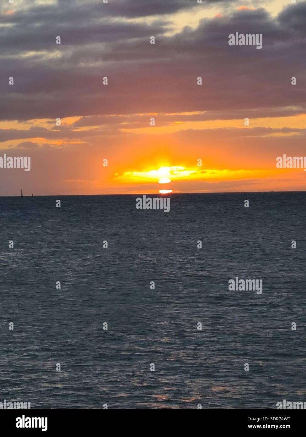 EDITORIAL USE ONLY- KEY WEST FLORIDA:  A vibrant tropical sunset over the Gulf, captured from Key West, Florida. The bright sun disk sinks toward a flat horizon line, framed by dramatic dark grey and orange-rimmed clouds. The deep blue water of the Florida Keys shows gentle surface ripples reflecting the intense golden hour glow. A distant maritime marker is visible on the horizon, emphasizing the vast coastal seascape. - Smartphone Captured Stock Image