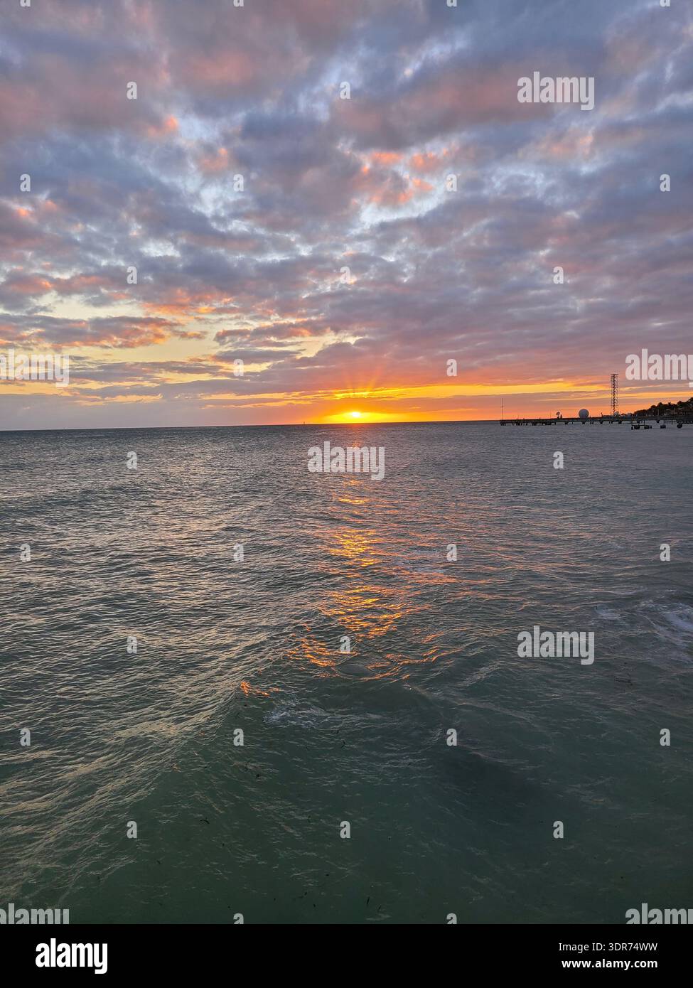 EDITORIAL USE ONLY- KEY WEST FLORIDA:  Vibrant sunset over a calm ocean horizon with scattered altocumulus clouds reflecting orange and pink light. A coastal pier and silhouette of a communications tower are visible in the distance. High-angle view from a beach or boardwalk. - Smartphone Captured Stock Image