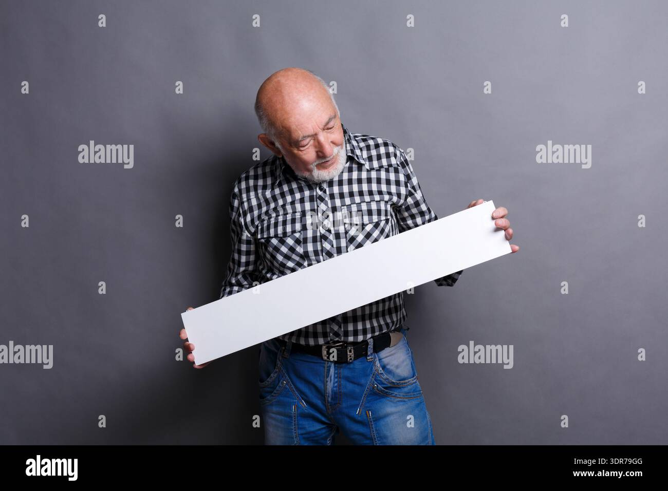 Older Man Holds Blank Sign While Standing Against Grey Background in Indoor Setting Stock Photo