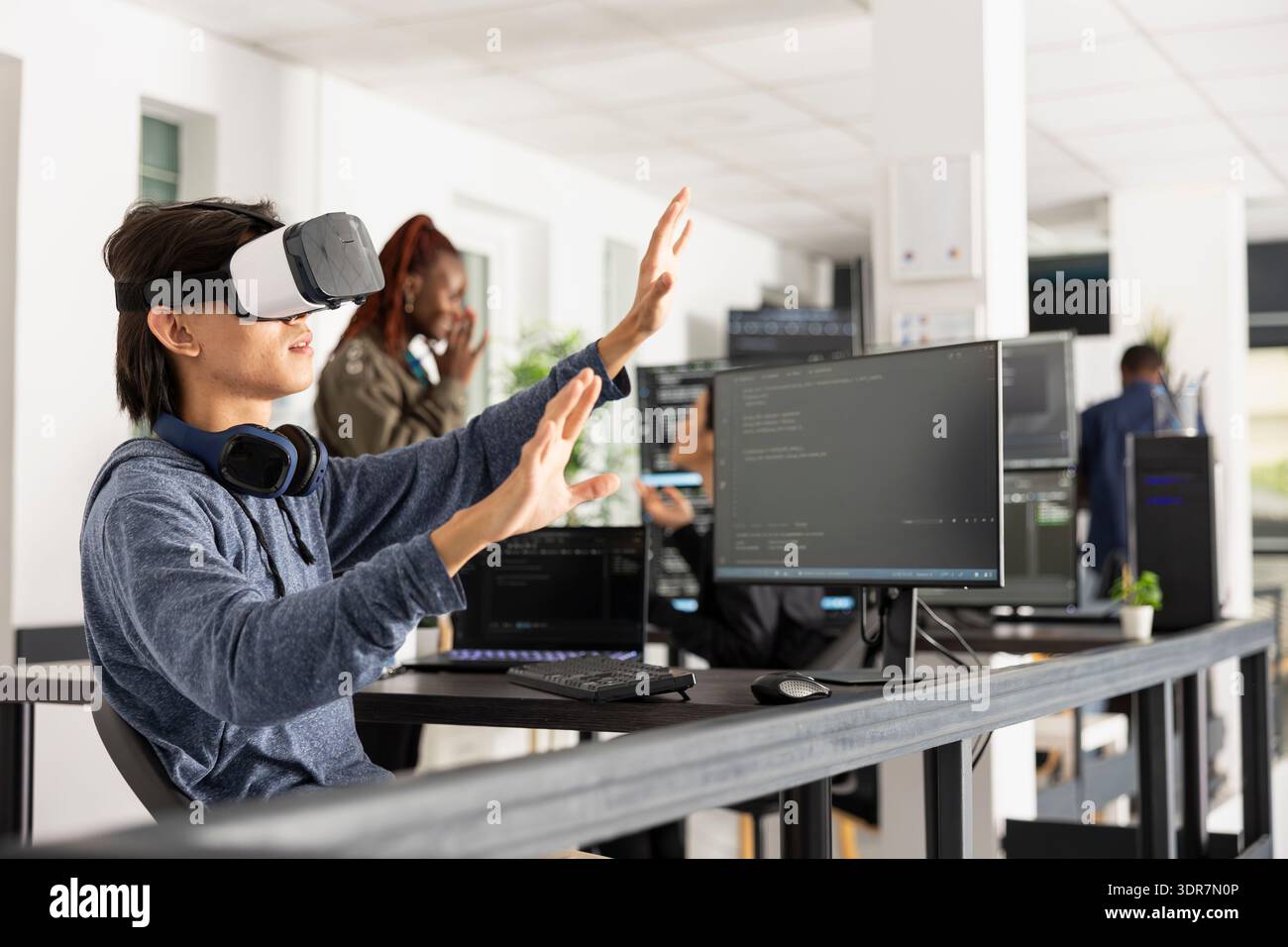 Male programmer uses VR glasses, interacting with 3D interface and analyzing coding scripts. Young male adult working at innovative company, exploring virtual technology for software development. Stock Photo