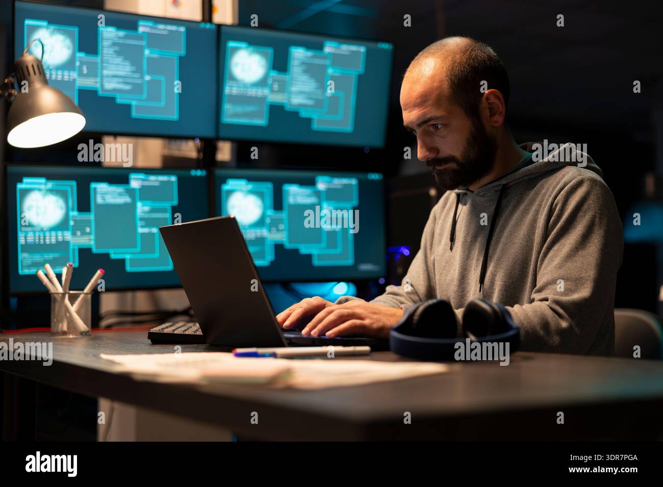Dedicated male programmer in sweatshirt reviews system data and writes coding scripts on laptop. Focused developer improving cloud computing protection to prevent cybercrime during night shifts. Stock Photo