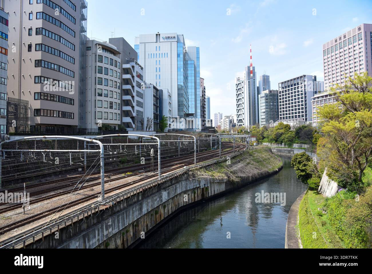 Wide view of multilane Tokyo rail infrastructure alongside a straight urban canal, flanked by office buildings and a red-white antenna mast. Stock Photo