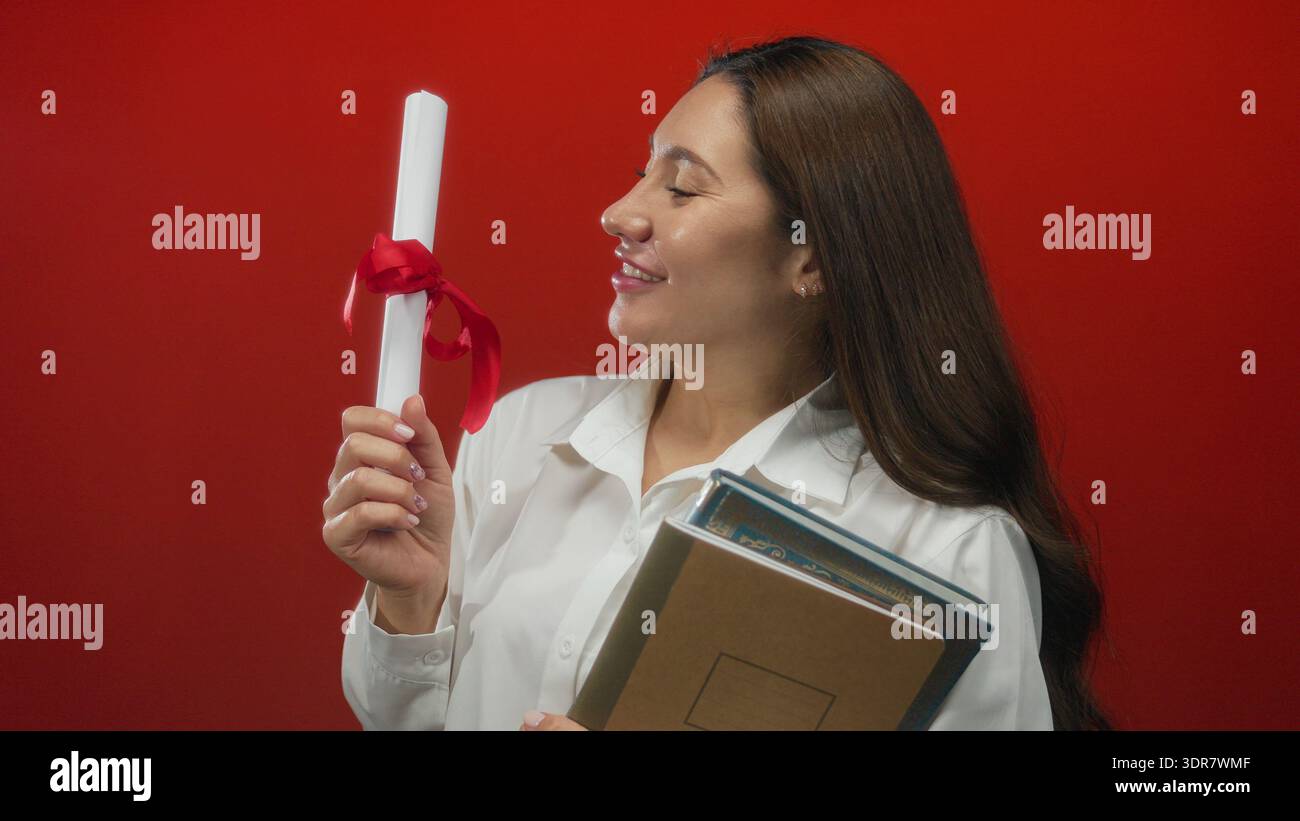Young hispanic woman holds diploma with red ribbon and stack of books in studio; proud achievement. Stock Photo