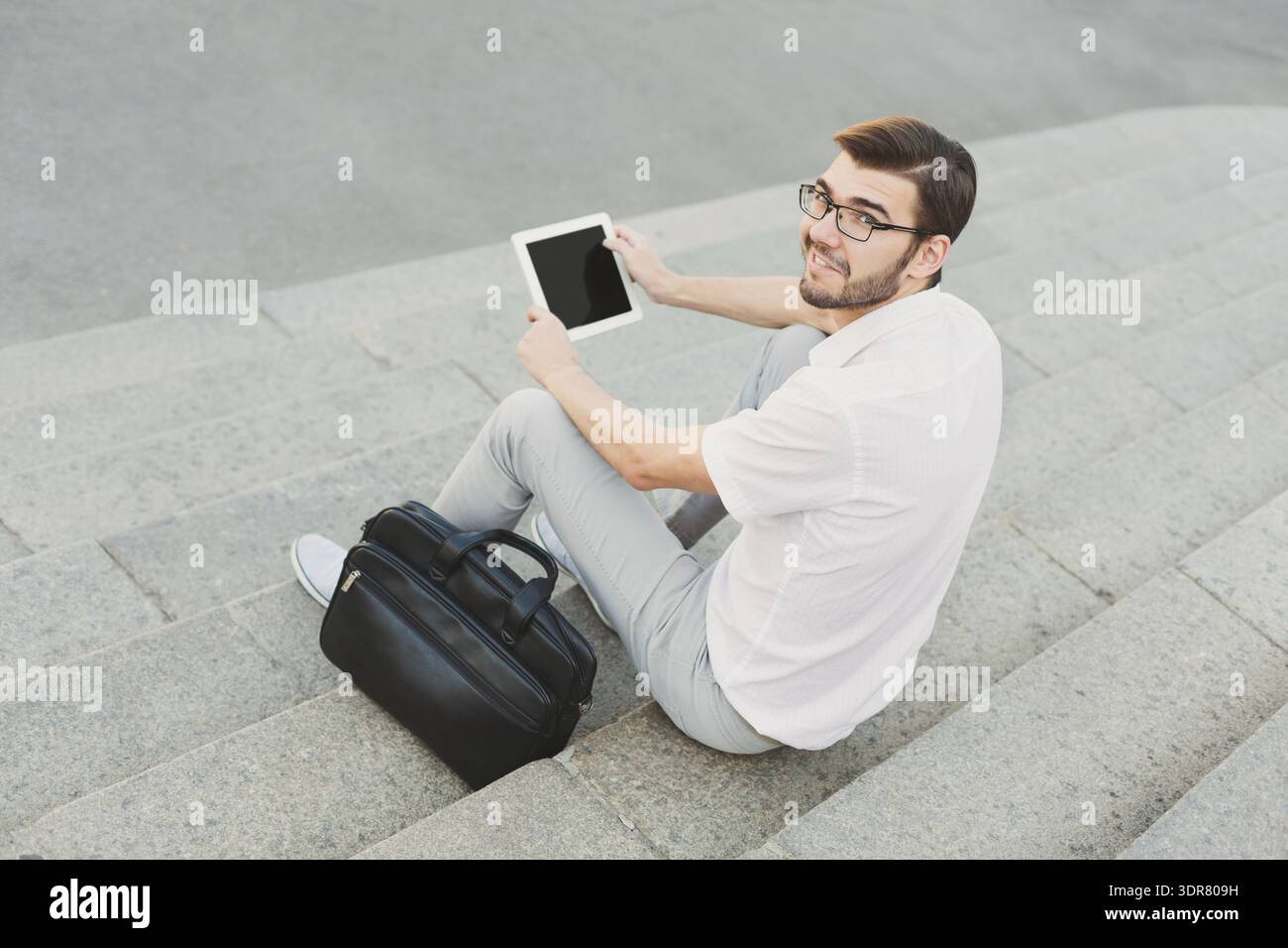 Man Uses Tablet While Sitting on Steps With Bag Beside Him Stock Photo