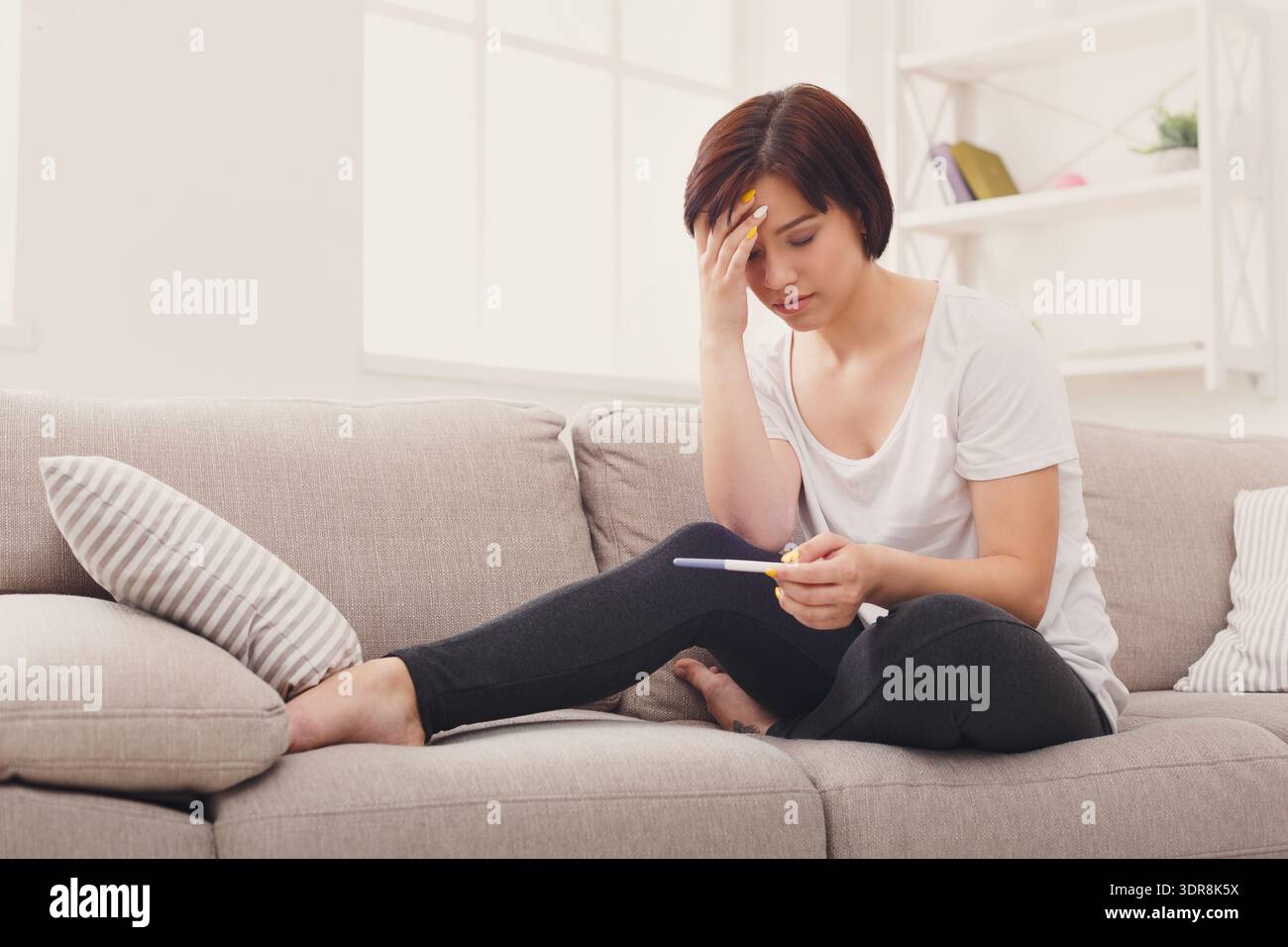 Woman Looks Worried While Sitting on Couch With Pregnancy Test in Hand at Home in Daylight Stock Photo