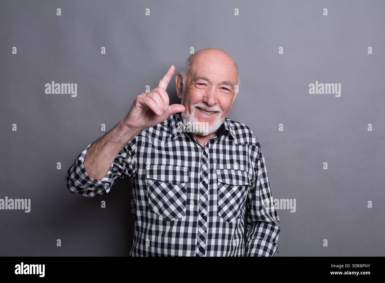 Elderly Man With Gray Beard Smiles and Shows Hand Gesture During Indoor Activity Stock Photo