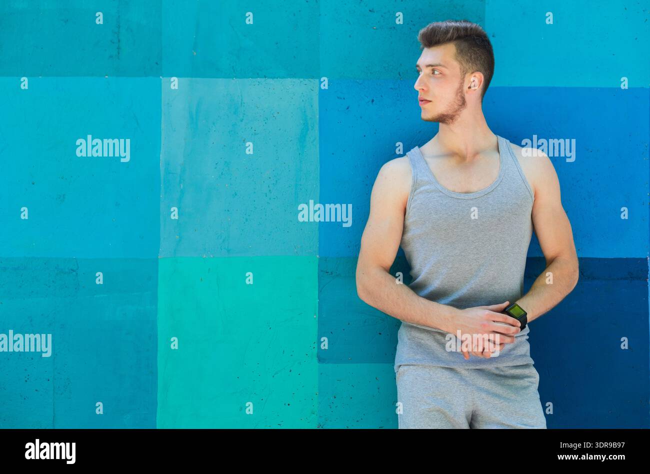 Young Man Stands Against a Blue-Green Wall While Holding a Watch During Daylight Hours Stock Photo