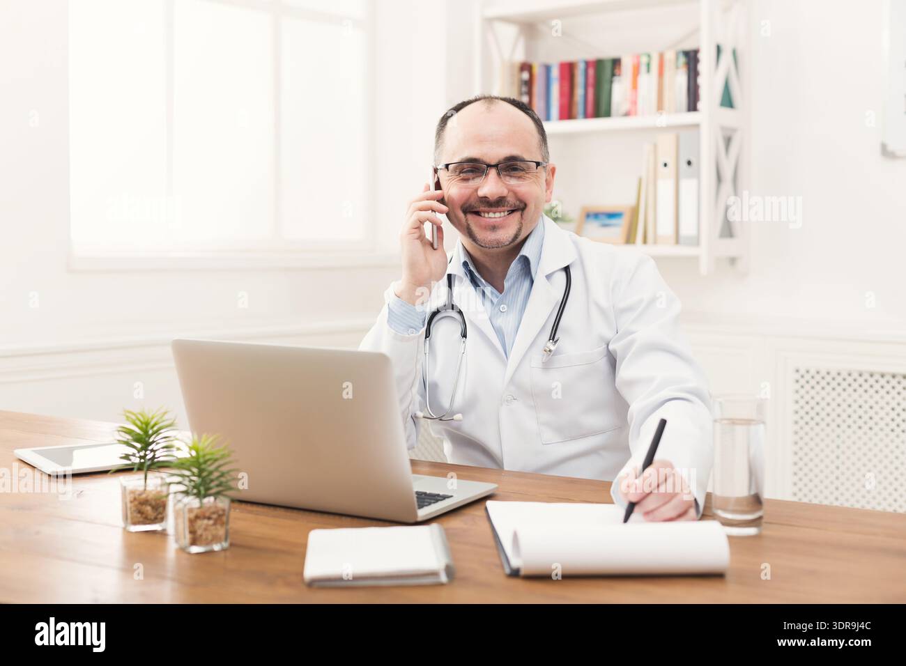 Doctor Talks on Phone While Taking Notes in Office During Working Hours Stock Photo