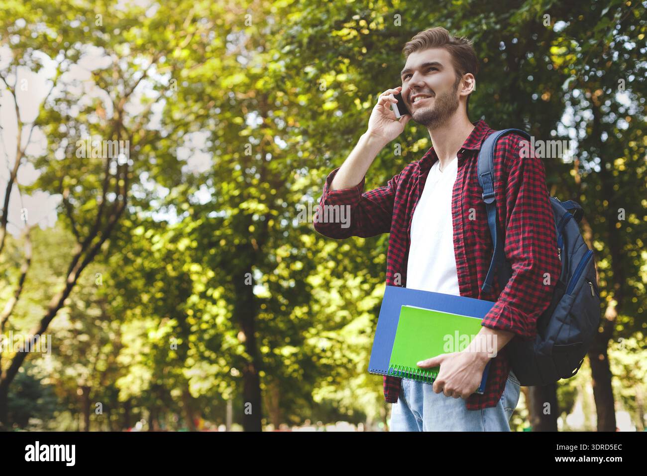 College Student Talks on Phone While Holding Notebooks in Park During Afternoon Stock Photo