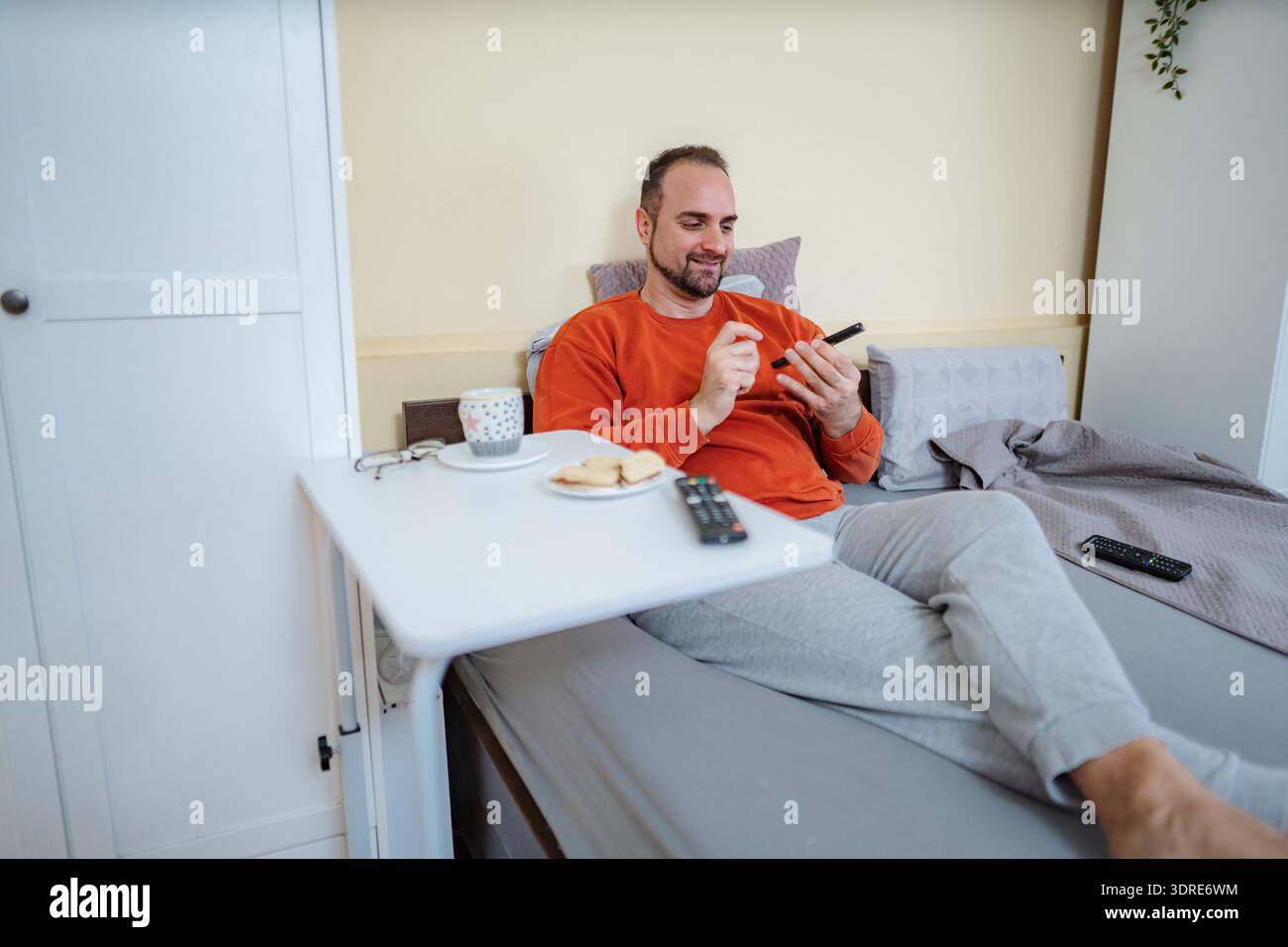 Man relaxing in bed enjoying breakfast and using smartphone Stock Photo