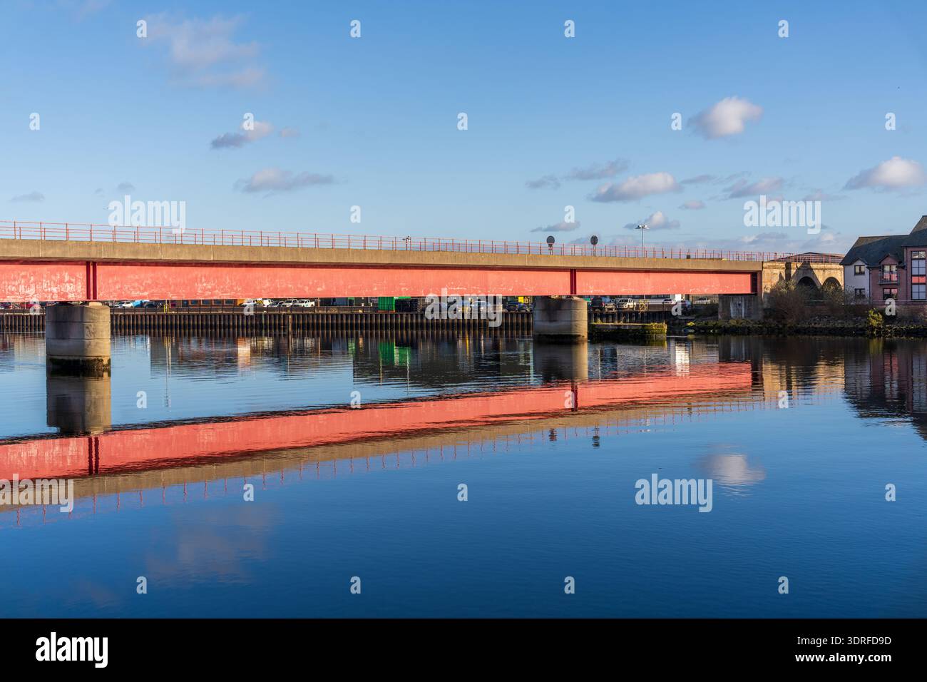 A long red bridge extends over the tranquil River Ness in Inverness, Scotland. Its vibrant color reflects perfectly on the smooth water surface under Stock Photo