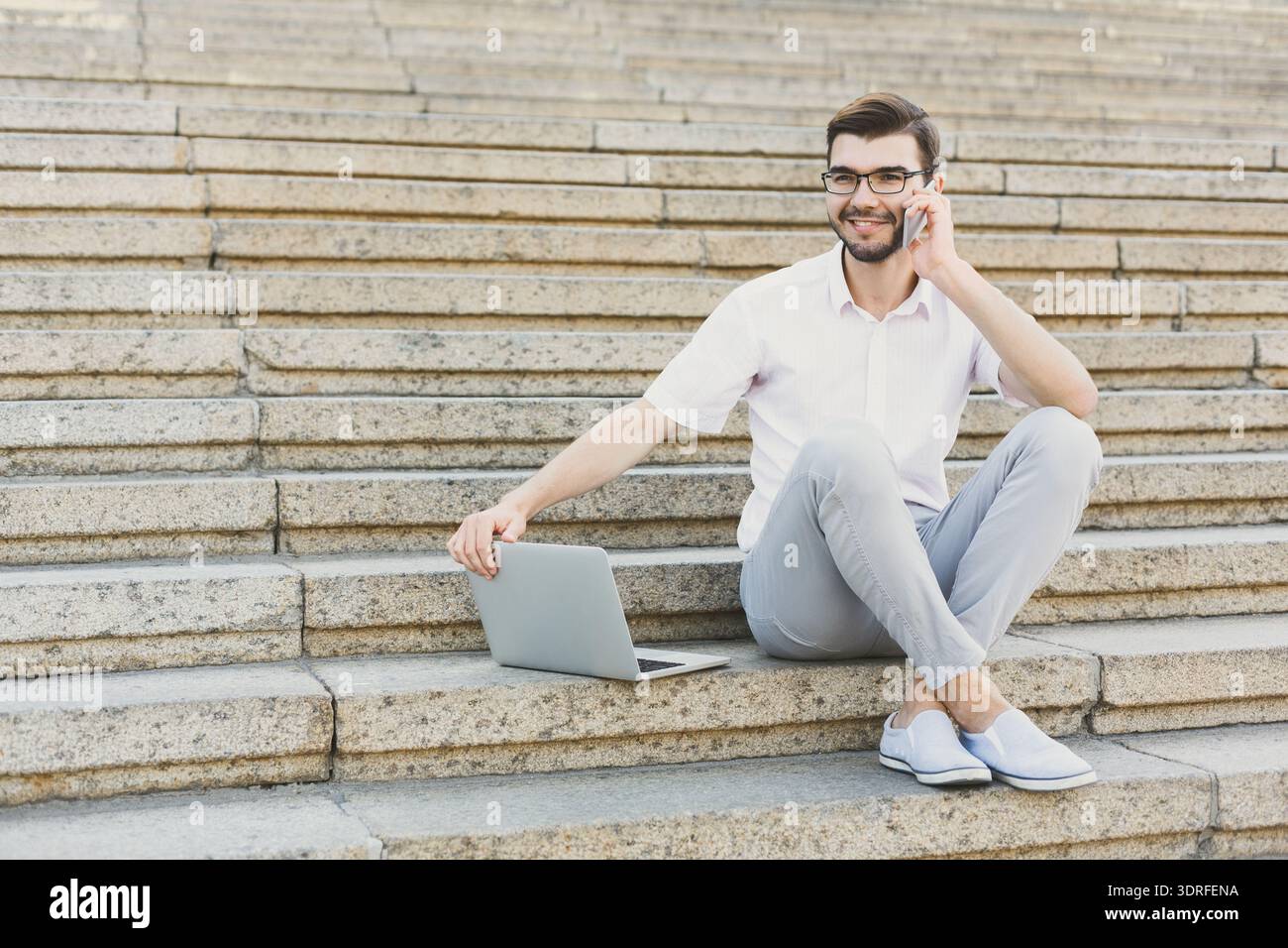 Young Man Sits on Stairs Using Laptop While Talking on Phone During Sunny Day Stock Photo