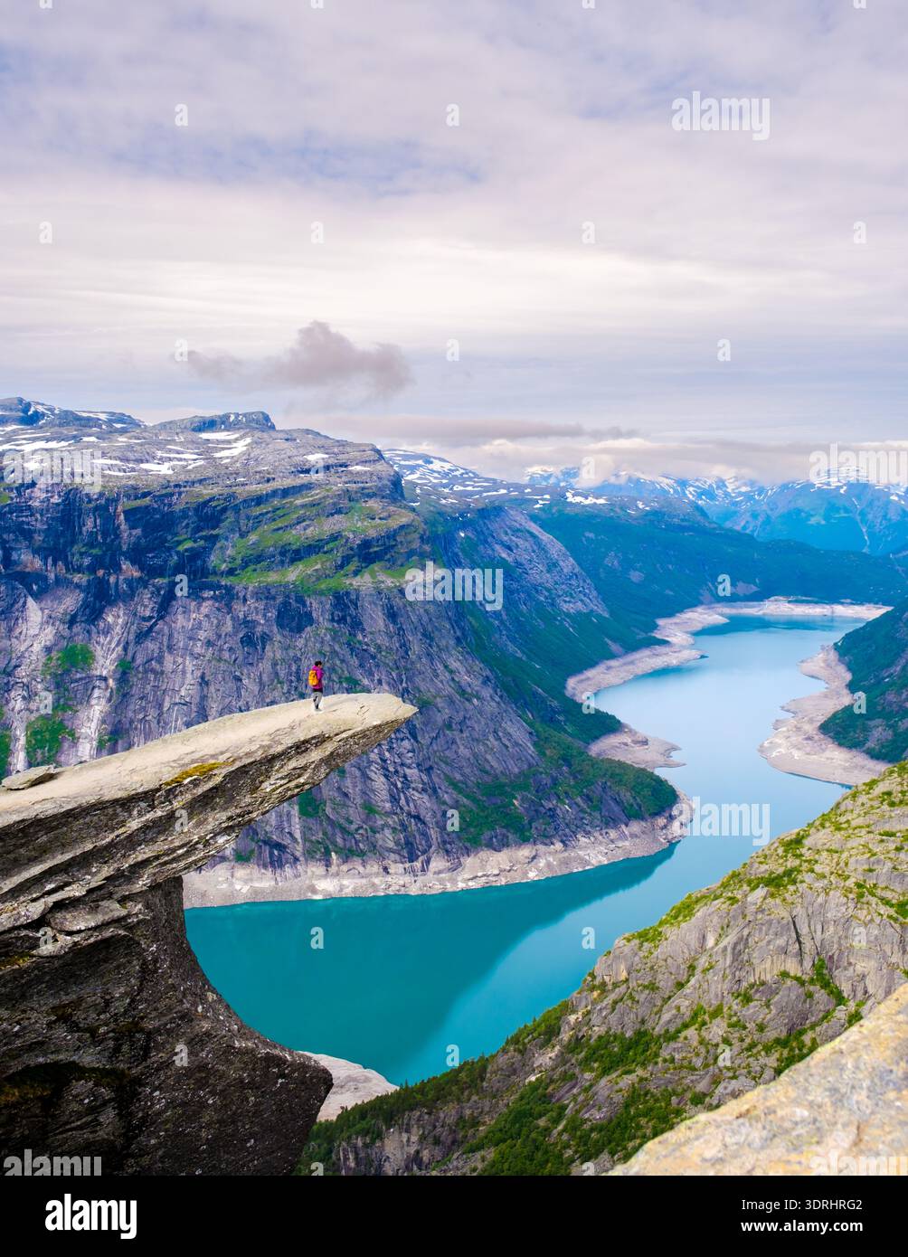 A lone hiker stands triumphantly on a dramatic cliff edge, gazing at the sparkling turquoise fjord below. Trolltunga, Norway Stock Photo