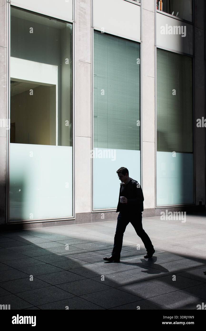 Silhouette of a businessman carrying coffee walking past the frosted glass facade of a Midtown Manhattan office building, New York City Stock Photo