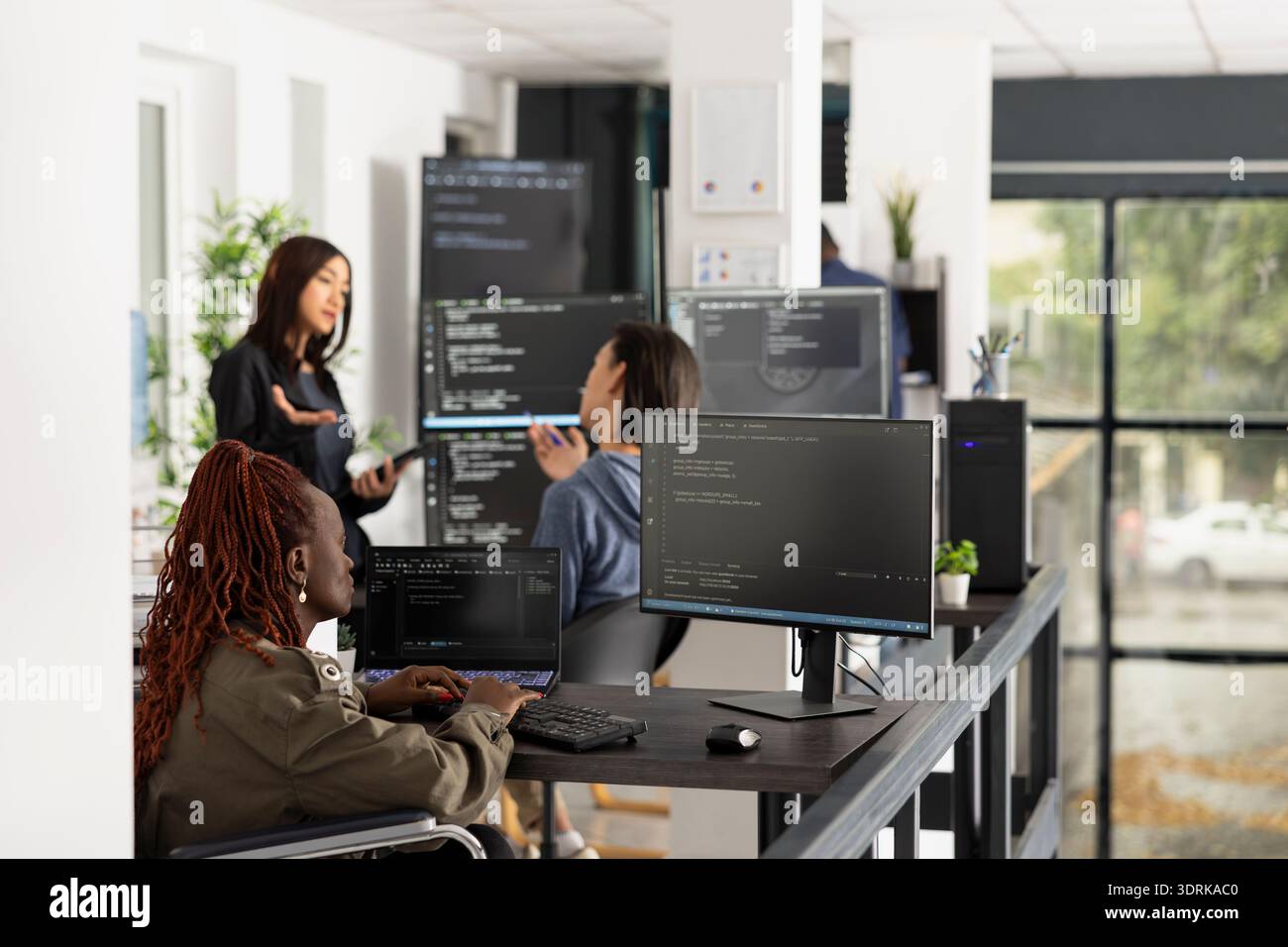 Black software developer in wheelchair studies frontend and backend code on computer screen. Focused professional demonstrates accessibility, problem solving and diversity within modern IT startup. Stock Photo