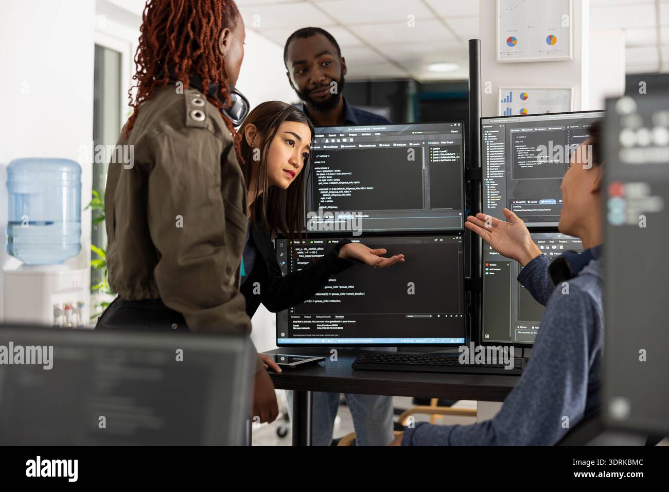 African American and Asian colleagues work side by side, analyzing source code and data on desktop monitors. Focused programmers collaborate to enhance web development projects in modern office. Stock Photo