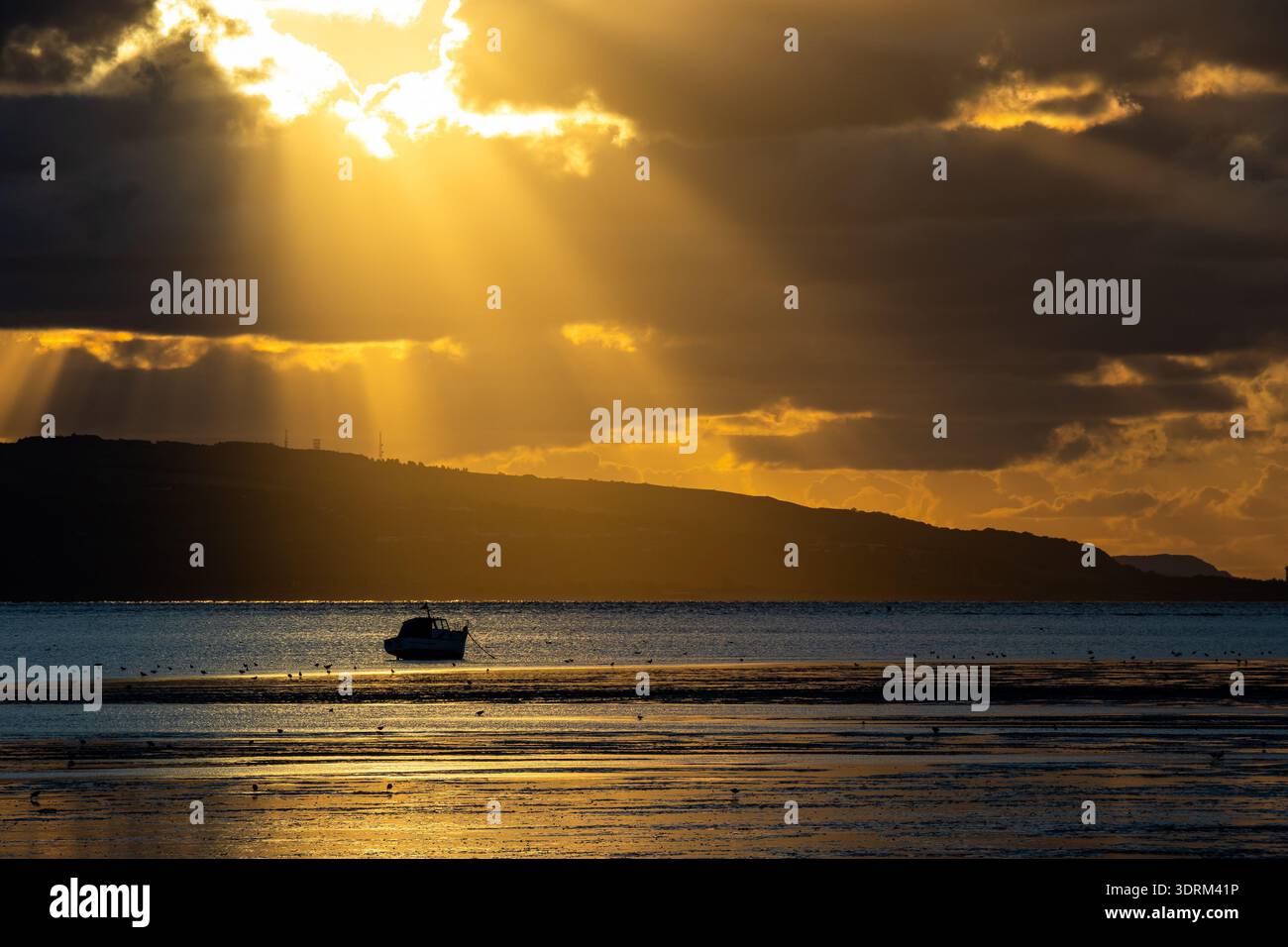 Golden sunlight rays breaking through dark clouds over a lone boat on the Dee Estuary Stock Photo