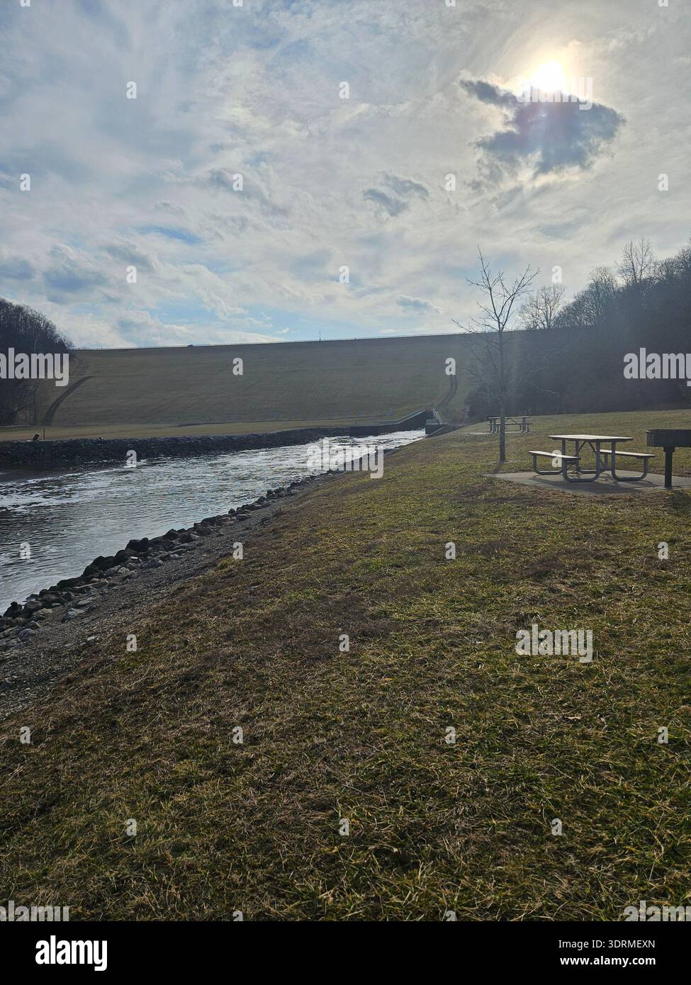 EDITORIAL USE ONLY- EAST FORK STATE PARK BETHEL OHIO:  : A view of the picnic area and tailwater below the William H. Harsha Lake dam at East Fork State Park, Bethel, Ohio. Empty picnic tables and park grills sit on a grassy bank beside the flowing water, with the towering earthen spillway rising against a cloudy, sunlit sky. - Smartphone Captured Stock Image