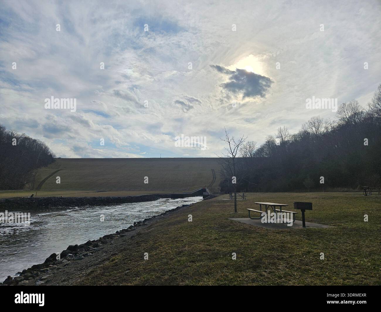 EDITORIAL USE ONLY- EAST FORK STATE PARK BETHEL OHIO: : A view of the massive earthen spillway and tailwater area at William H. Harsha Lake within East Fork State Park, Ohio. The scene features a riverside picnic area with tables and grills in the foreground, with the towering dam structure rising against a cloudy afternoon sky with a diffused sun. - Smartphone Captured Stock Image