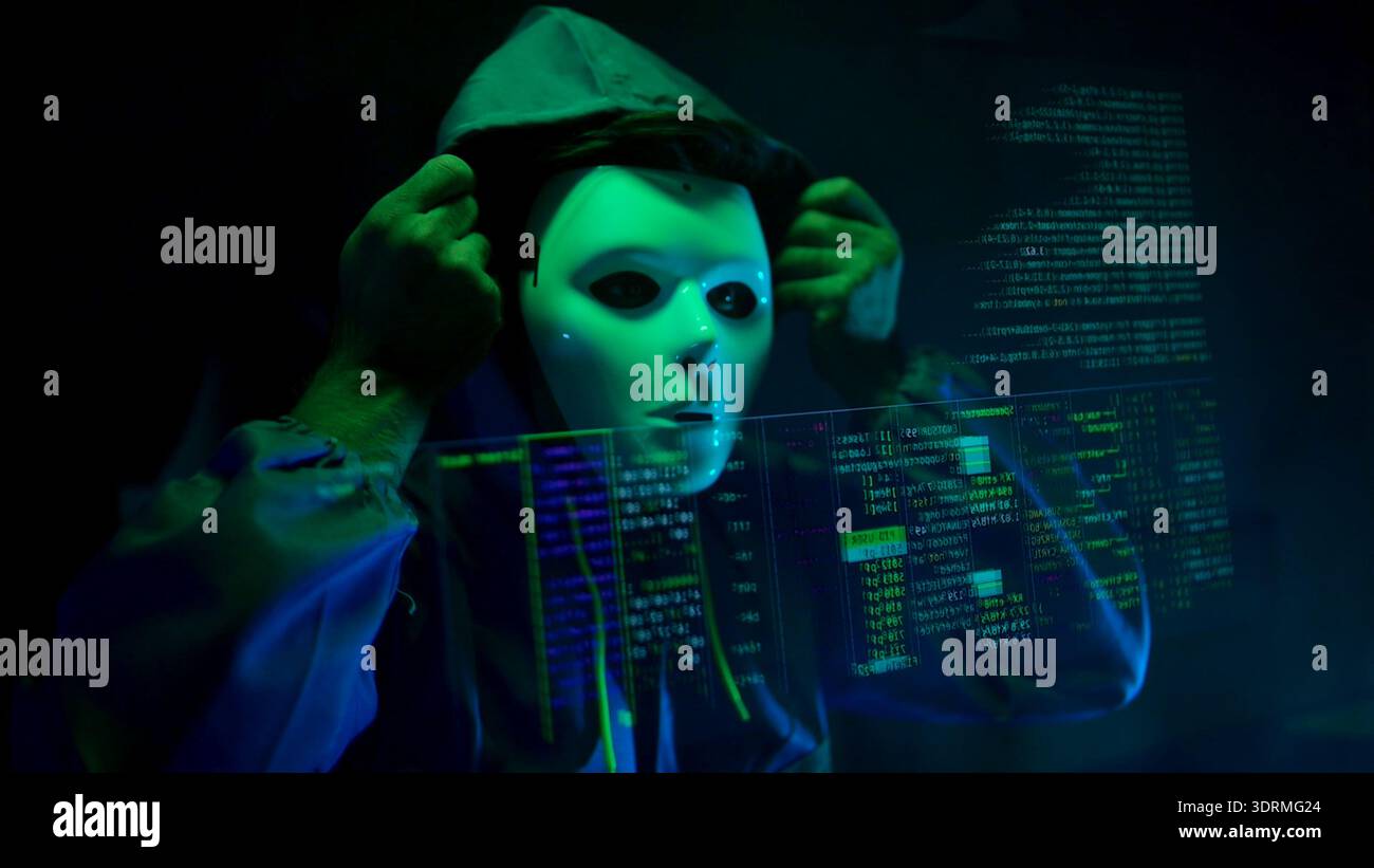 Anonymous hacker wearing a mask and hoodie interacting with digital data in a dark room Stock Photo