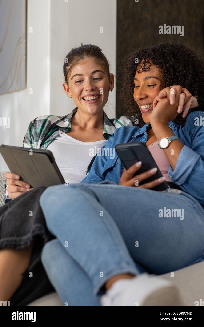 Diverse female friends reclining on light-colored couch in living room using tablet and smartphone. Casual, friendship, relaxati Stock Photo