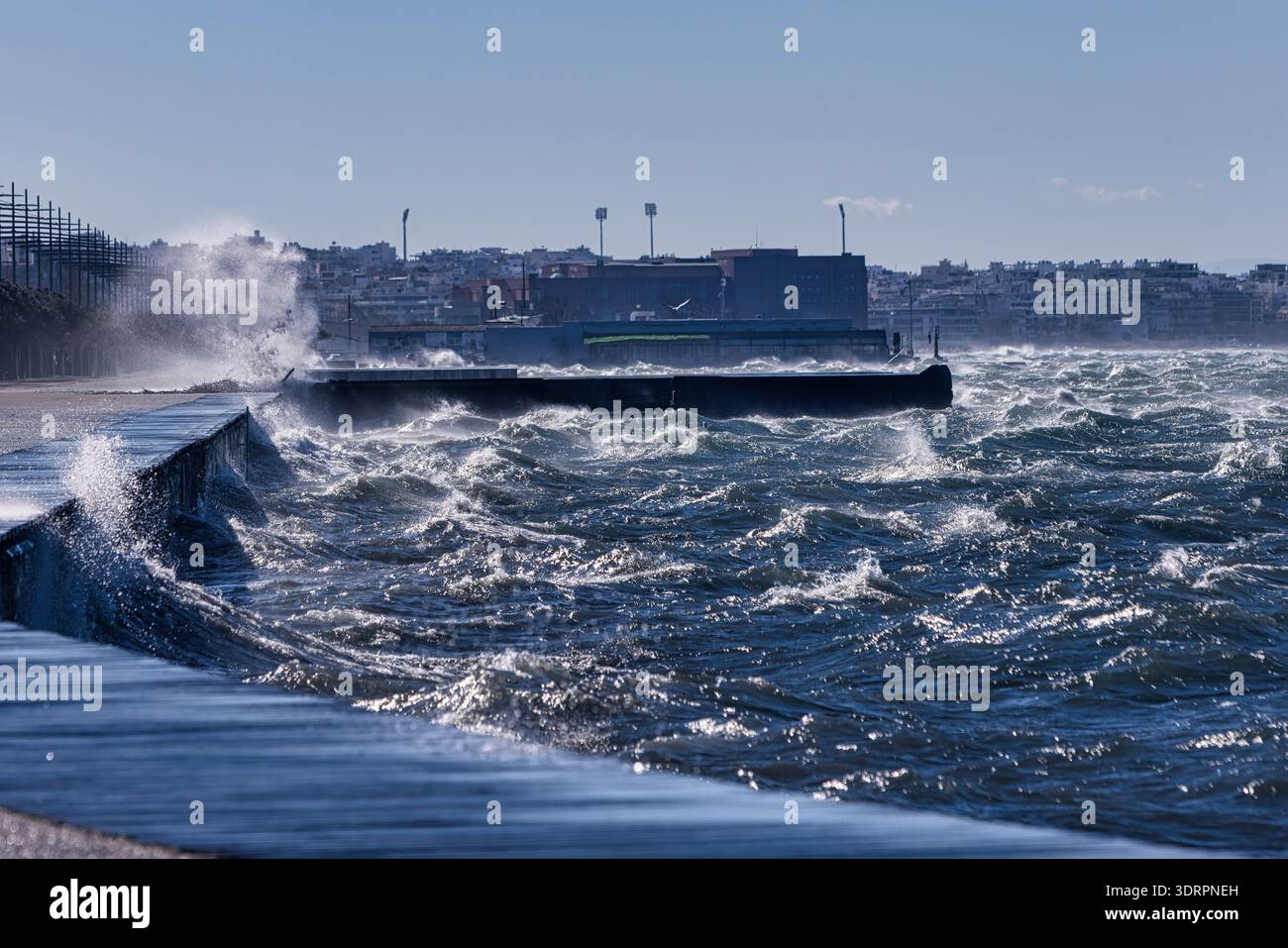 Powerful waves crash against the waterfront promenade in Thessaloniki, creating dramatic splashes along the curved shoreline. A person and a dog walk Stock Photo