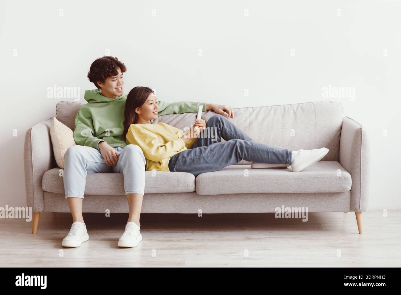 Affectionate young Asian couple using smartphone, relaxing on couch together at home, free space Stock Photo