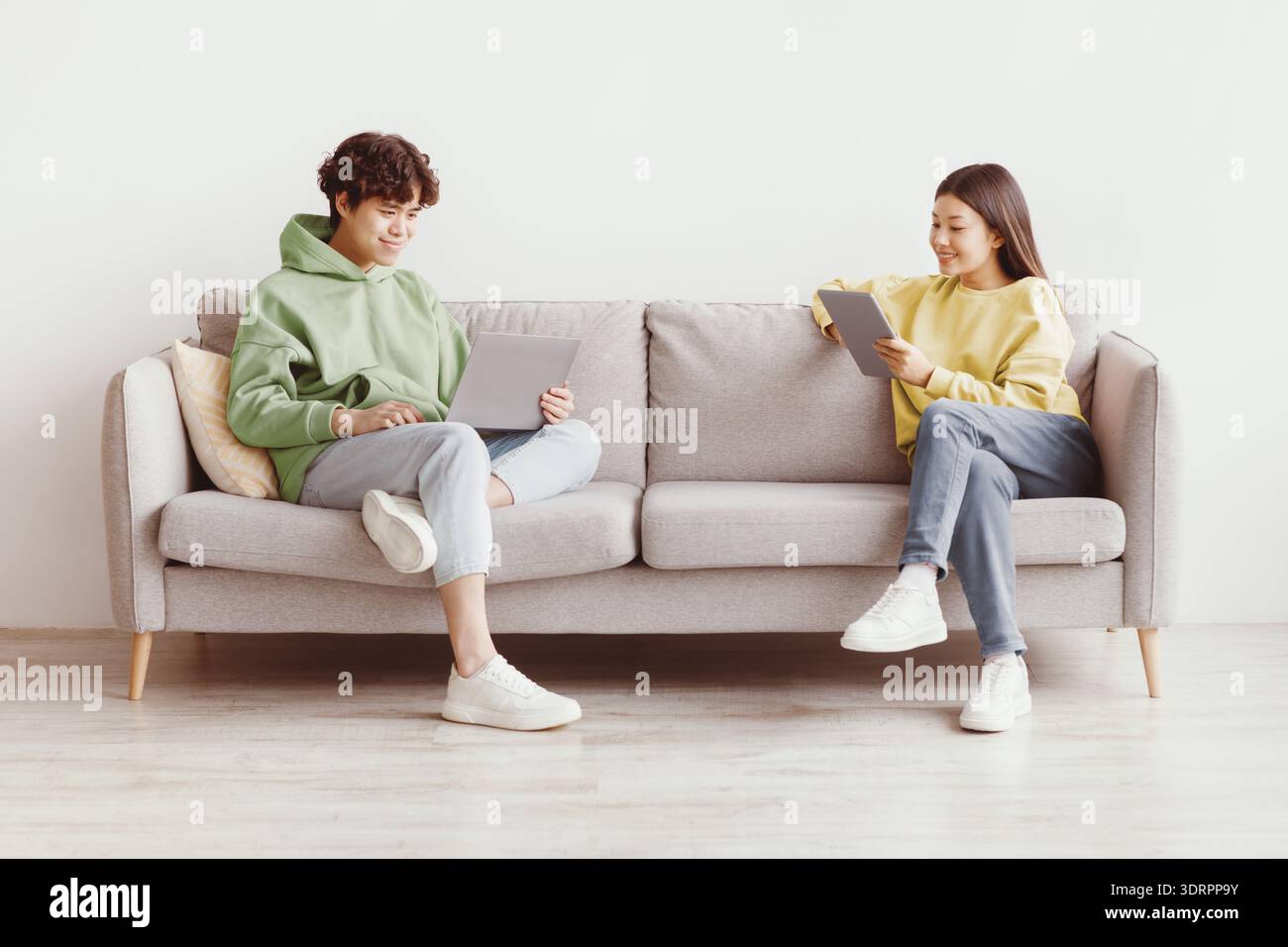 Handsome young Asian guy and his girlfriend using modern gadgets while sitting on couch at home, copy space Stock Photo