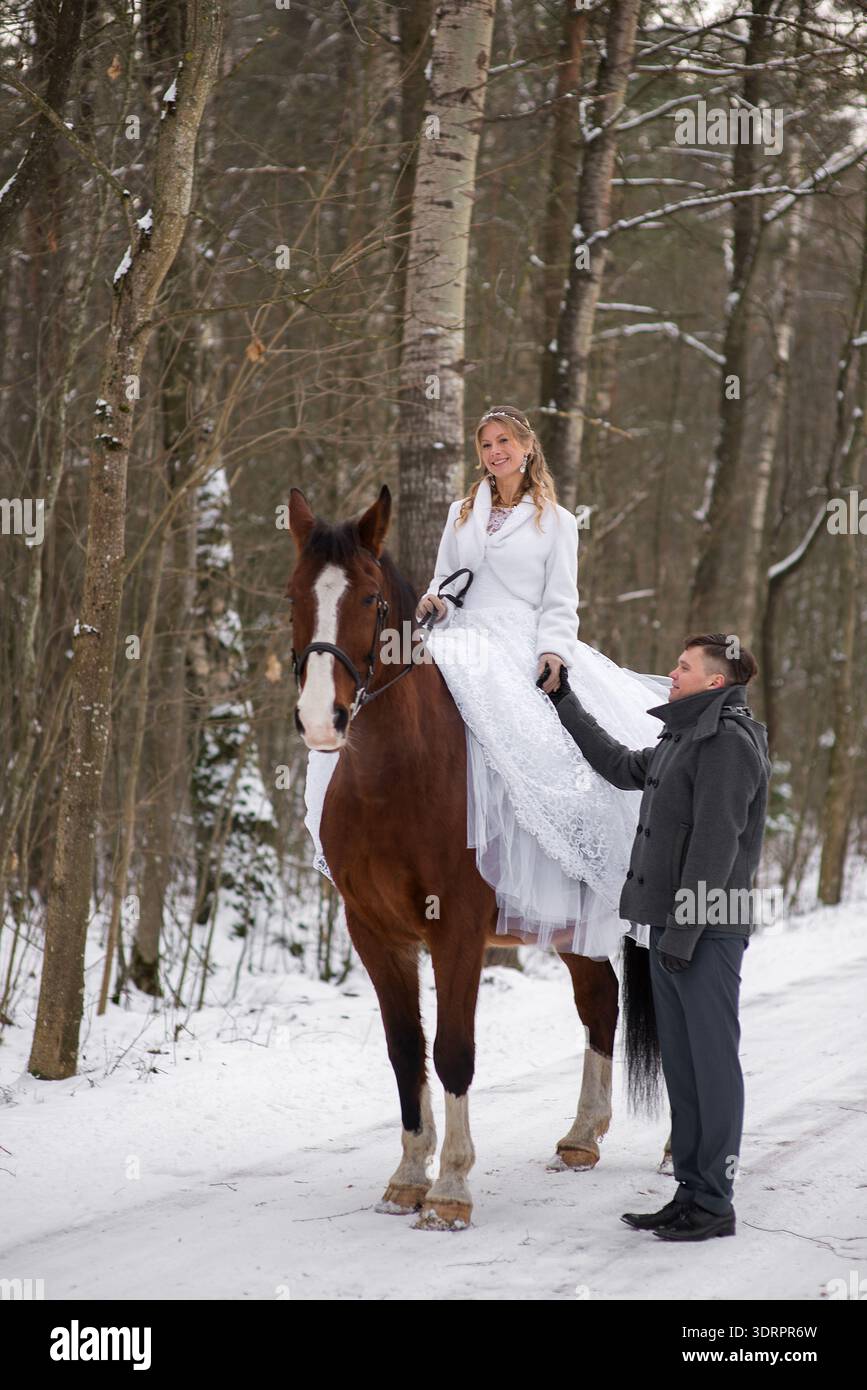 Groom stands beside a brown horse while the bride sits on the saddle in a white wedding dress, smiling gently during a calm winter photoshoot in the f Stock Photo