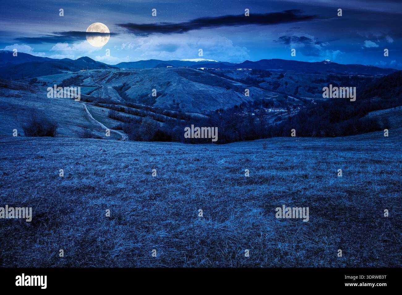 rural landscape with village in valley at night. outdoor adventures in countryside with rolling hills and snowy mountain in early spring in full moon Stock Photo