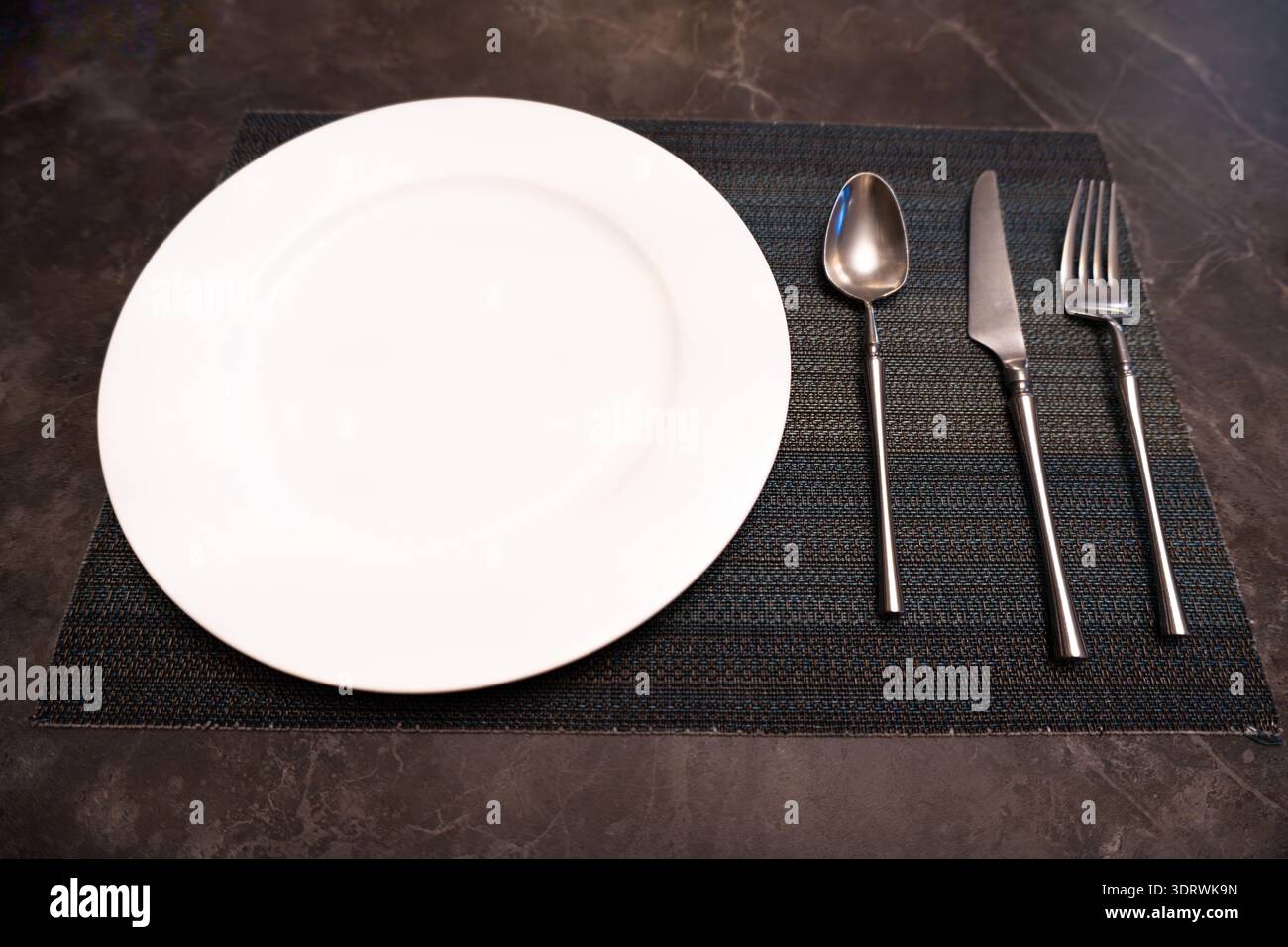 Empty white dinner plate with silverware on dark textured placemat, minimalist table setting Stock Photo