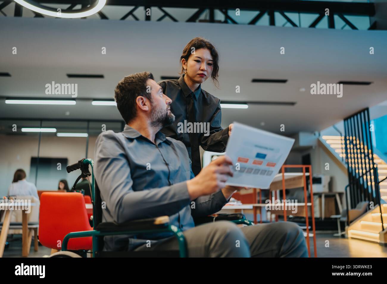 Team collaboration in a modern office as a woman assists man in a wheelchair with documents Stock Photo