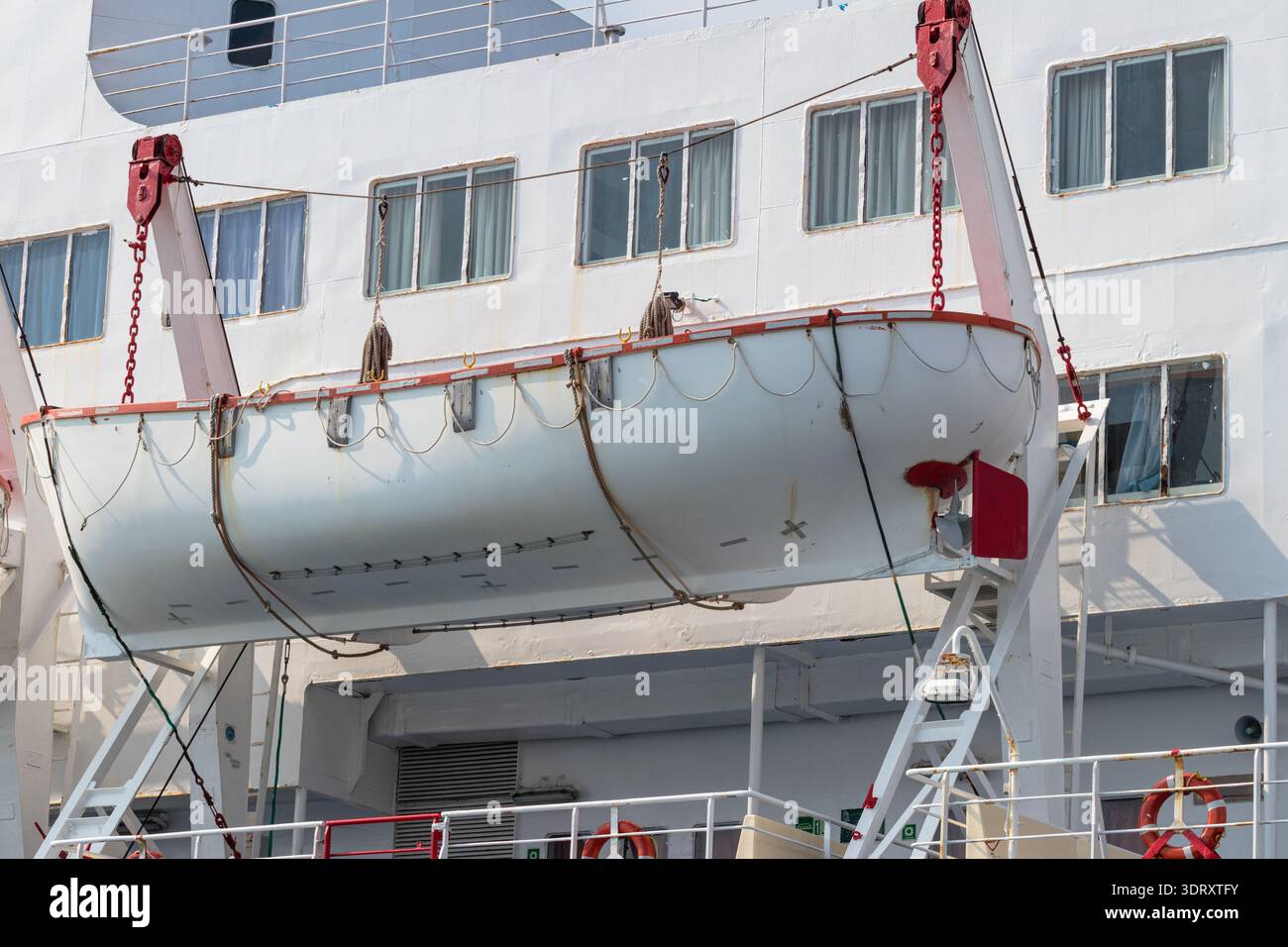 A white lifeboat suspended on davits beside the hull of a cruise ship ...