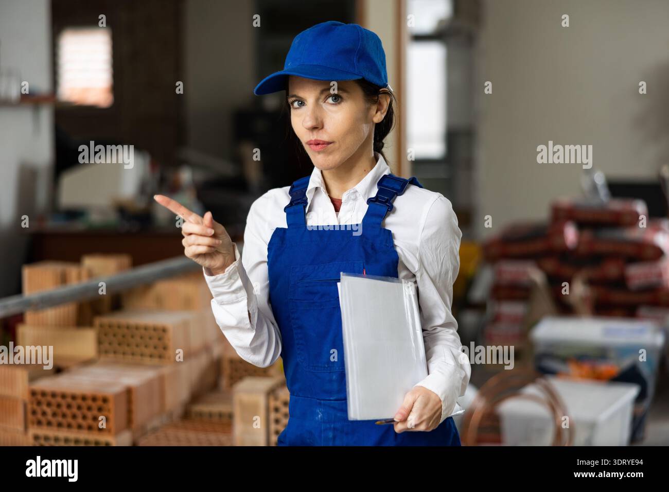 Female builder wearing overalls and cap posing at construction site, looking at camera Stock Photo
