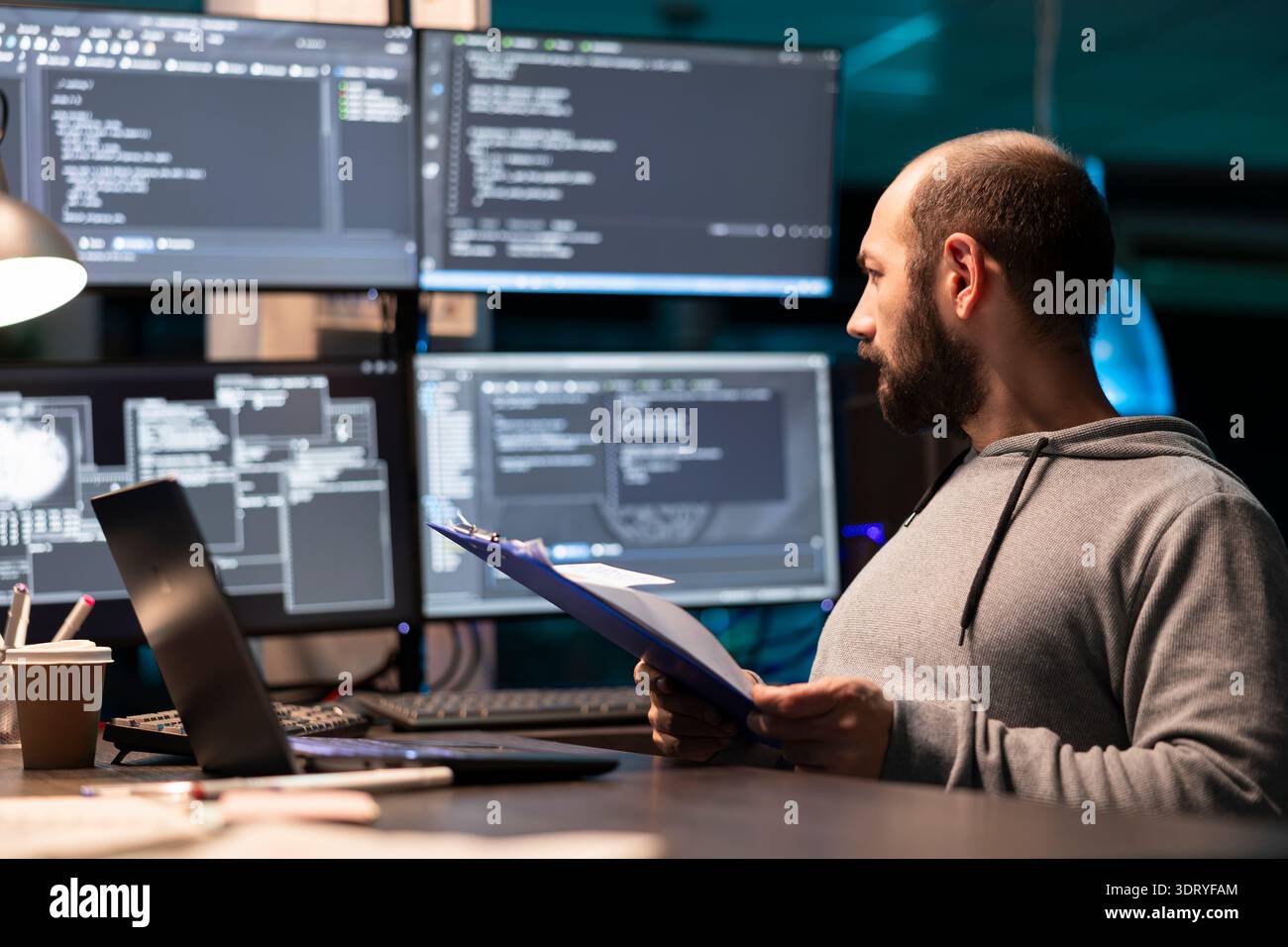 Programmer examines paperwork with serious expression and compares notes to code displayed on surrounding monitors. Developer with clipboard glancing at large displays of data, ensuring accuracy. Stock Photo