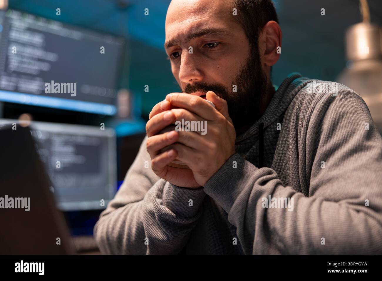 Tired software engineer warms his hands on hot coffee cup while thinking through late night debugging tasks. Concerned programmer stares at computer display, worried about system issues and deadlines. Stock Photo