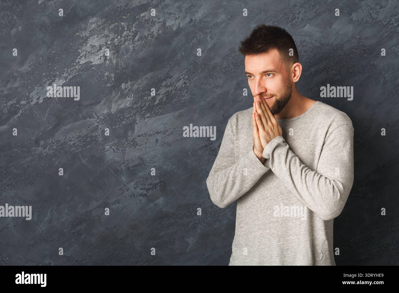 Young Man Stands With Hands Clasped Together in a Thoughtful Pose Against a Gray Wall Backdrop Stock Photo