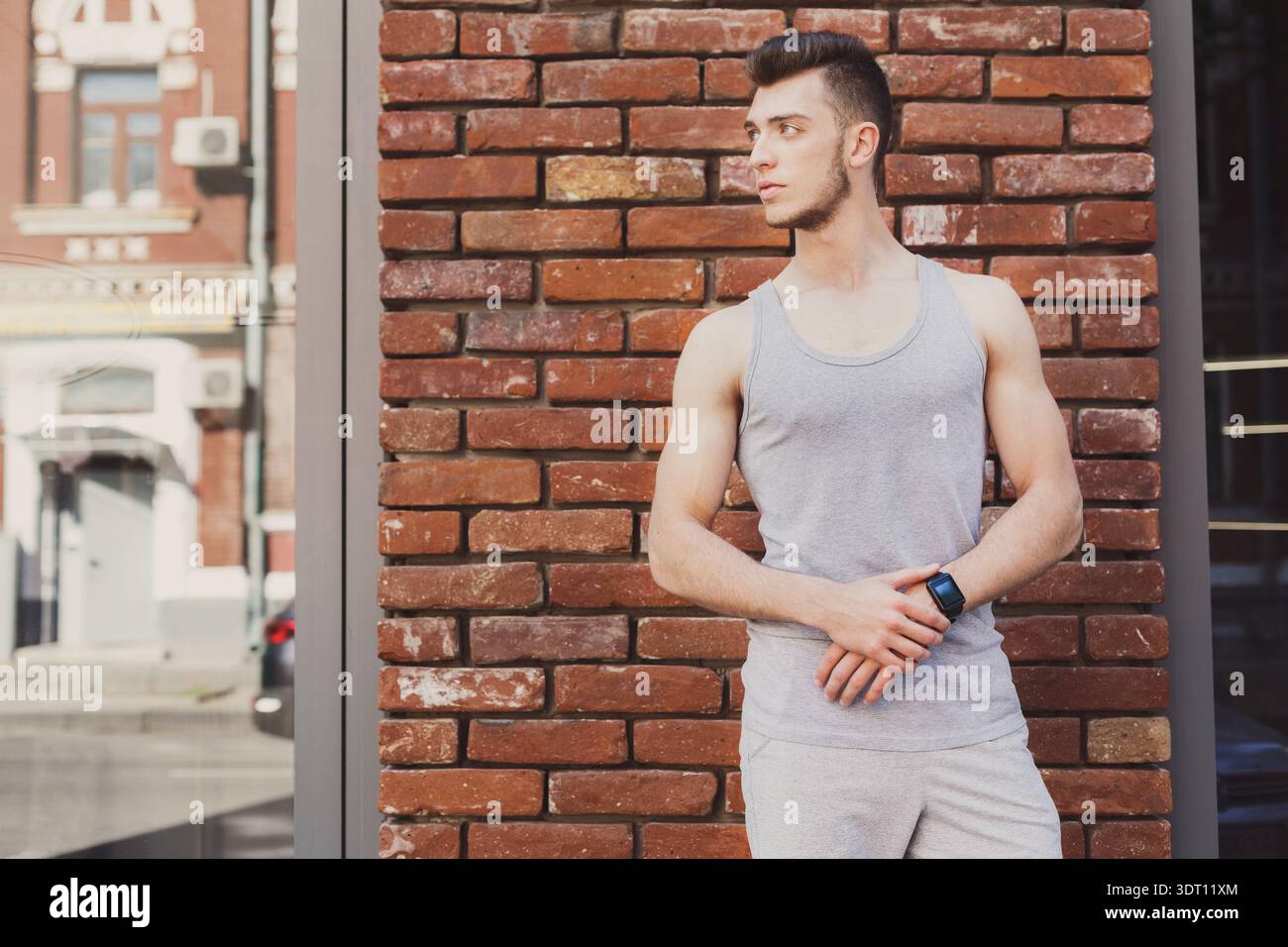 Young Man Stands by Brick Wall in Urban Area While Checking His Watch in the Afternoon Stock Photo