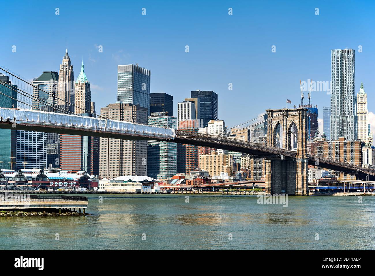 Brooklyn Bridge stands tall above water while Manhattan skyline showcases buildings on a clear day in New York City. Stock Photo