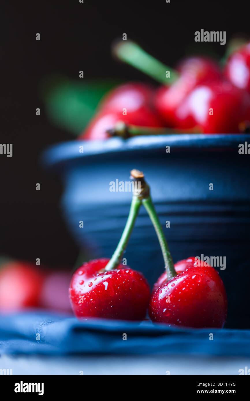Two sweet cherries in front of dark bowl, selective focus composition. Fresh red summer fruit with stems, minimal food styling Stock Photo