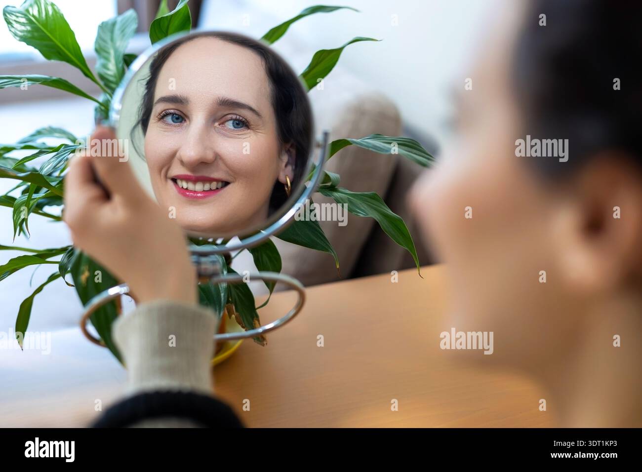 Smiling woman looking in mirror at home. Happy young woman checks her face in a small tabletop mirror, natural makeup and bright daylight with green Stock Photo