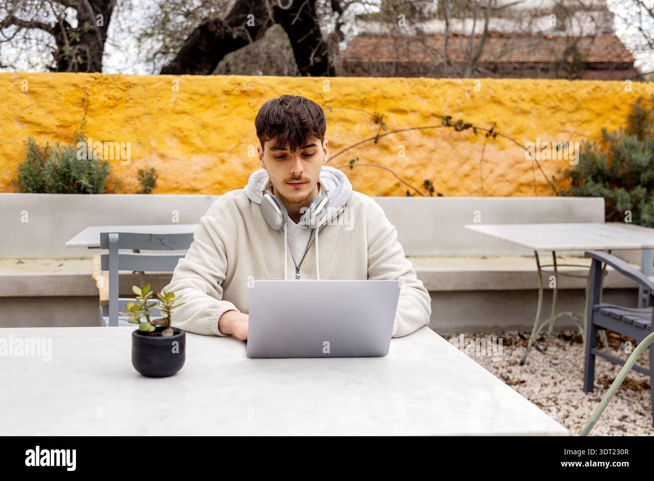 Young man focused on his laptop while working outdoors. Dressed casually with headphones around his neck, he's tapping into technology to complete fre Stock Photo