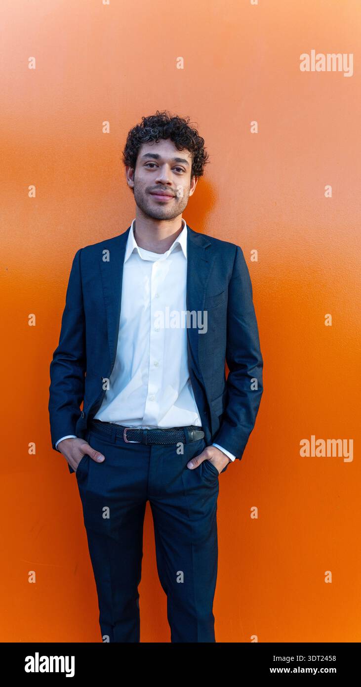 A confident young businessman in a sharp navy suit stands against a vibrant orange wall. His relaxed posture and friendly demeanor convey professional Stock Photo