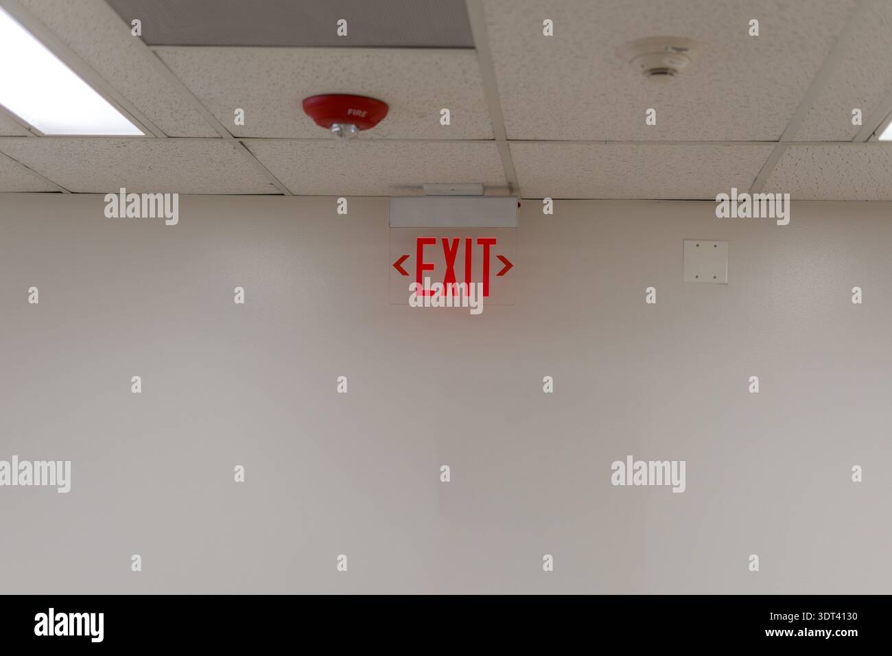 A horizontal shot of a glowing red EXIT sign mounted on a white wall below a drop ceiling. The image also captures a red fire alarm strobe and a smoke Stock Photo